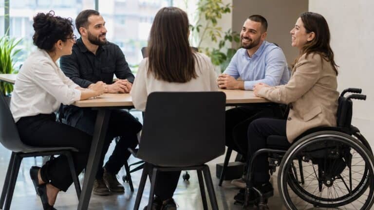 חמש דמויות מגוונות יושבות סביב שולחן ישיבות במשרד ומנהלות שיחה.