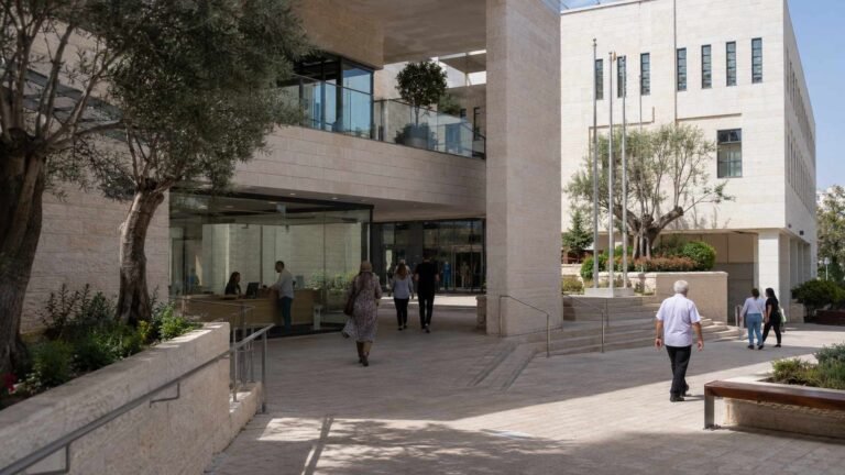 אנשים הולכים ברחבה מרוצפת בחצר של מתקן ציבורי מודרני הבנוי מאבן בהירה.